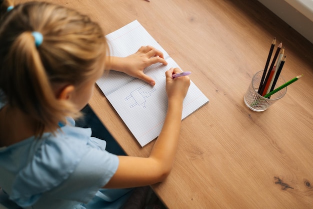 Высокоугольный вид сбоку на неузнаваемую маленькую девочку младшего возраста, рисующую ручкой, делающую картинку сидящей за столом в светлой детской комнате у окна
