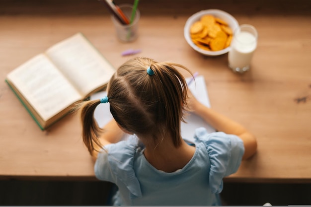 クッキーとミルクと一緒に家のテーブルに座って学習している小学生の女の子のハイアングルビュー