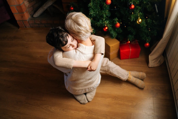 Highangle view of happy child son hugging loving young mother near Christmas tree decorated with balls and cozy fireplace