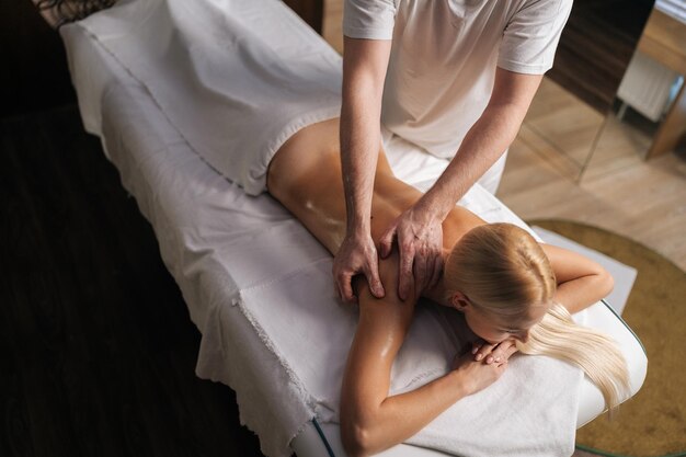 Фото Высокоугольный вид неузнаваемого массажиста-мужчины, делающего массаж спины и плеч блондинке-клиентке с красивой кожей, лежащей в спа-салоне