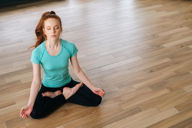 Highangle view of serene young woman in sportswear meditating in lotus position sitting on floor with closed eyes at home enjoying meditation