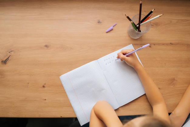テーブルに座ってペンでコピーブックに宿題を書いている認識できない小学生の女の子のハイアングルビュー