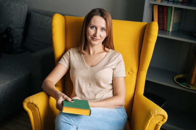 Highangle weergave van mooie blonde vrouw rustend in comfortabele gele stoel met papieren boek in donkere kamer met modern interieur