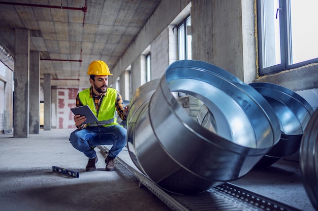 Operaio edile caucasico altamente motivato in abbigliamento da lavoro accovacciato accanto ai tubi di scappamento e controllando la loro qualità. nelle mani è tablet.