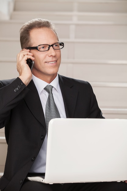 Hij heeft altijd contact. vrolijke volwassen man in formalwear die op de mobiele telefoon praat en computer gebruikt terwijl hij op de trap zit