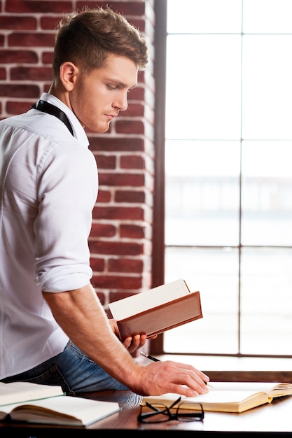 Hij houdt van studeren. Knappe jonge man in overhemd en stropdas die iets in notitieblok schrijft terwijl hij op zijn werkplek zit