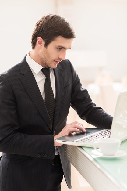 Hij is altijd verbonden. Zelfverzekerde jonge man in formele kleding die zijn laptop gebruikt terwijl hij aan de bar staat