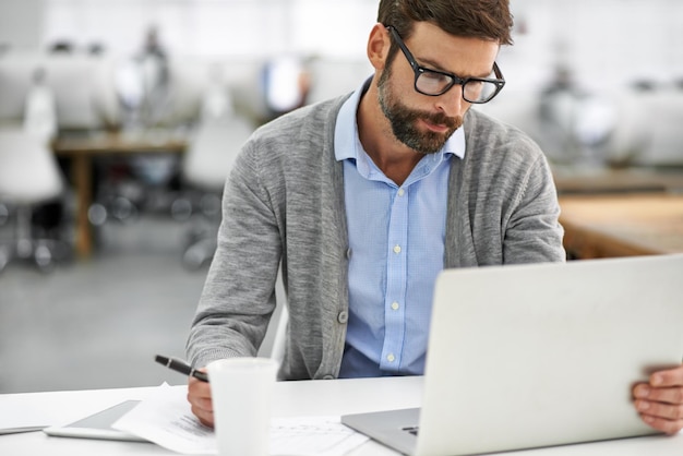 Hij is hard aan het werk op zijn laptop Een jonge zakenman die over papierwerk gaat terwijl hij op zijn laptop op kantoor werkt