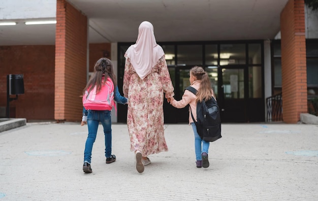 ヒジャブのイスラム教徒の母親は、学校の幸せなイスラム教徒の家族から2人の子供を手をつないで歩いています。