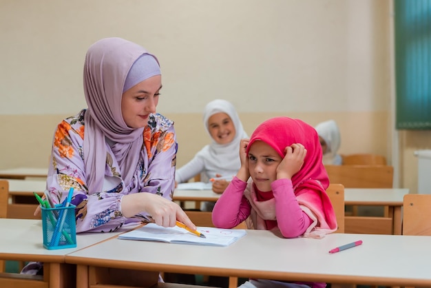 Hijab Muslim teacher helps confused and stressed school child to finish the lesson in the classroom.