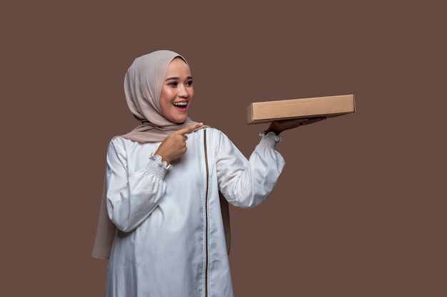 Foto hijab vrouw met een pizzadoos met één linkerhand en haar rechterhand wijzend terwijl ze lacht