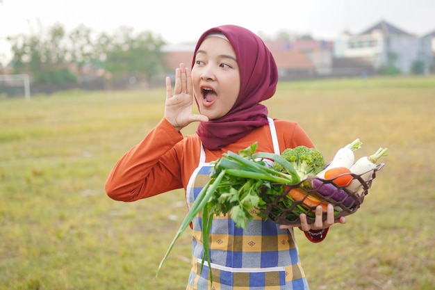 ヒジャブの女性はエプロンを着用し、野菜の入ったバスケットを保持してジェスチャーを叫ぶ