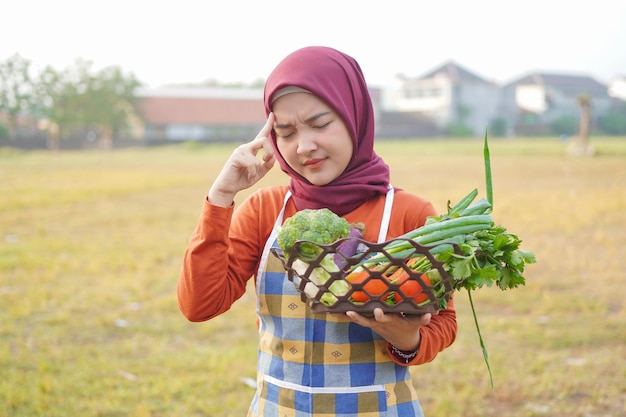 ヒジャブの女性はエプロンを着て野菜を持ち、考えるジェスチャーをする