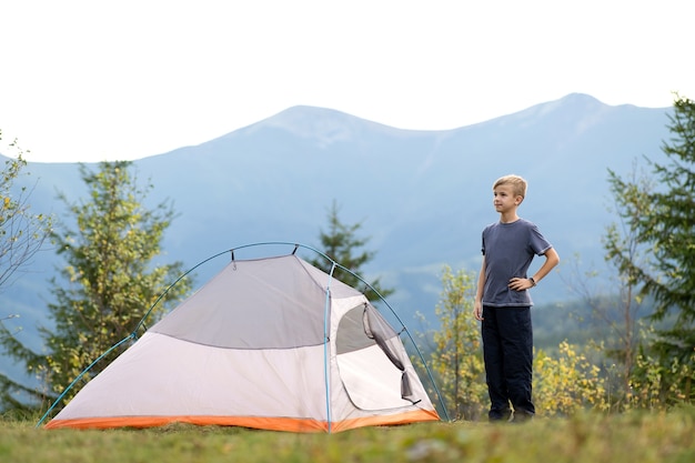 Hiker child boy resting near a tourist tent at mountain campsite enjoying view of beautiful summer nature.