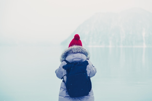 Hiker girl with backpack in down jacket and red cap standing near lake during snowfall in winter. Travel concept.