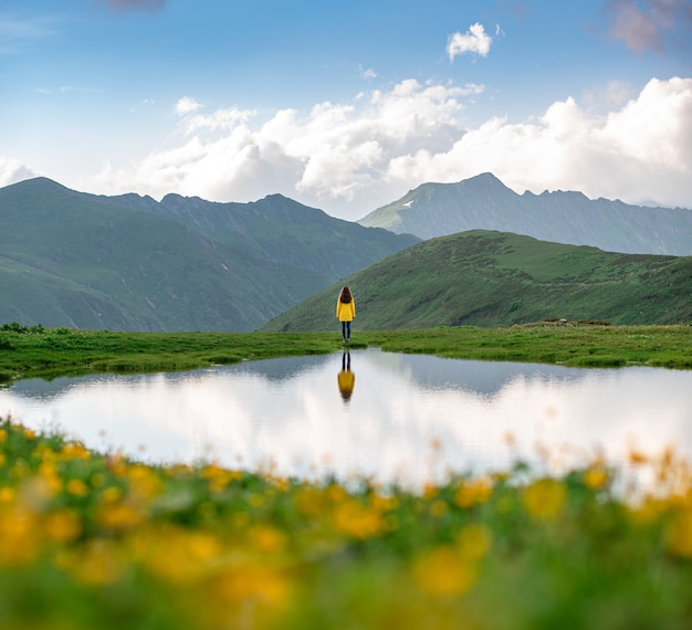 A hiker girl in a yellow jacket running on the shore of a lake among flowering meadows. beautiful reflection of a woman in the water of a mountain lake