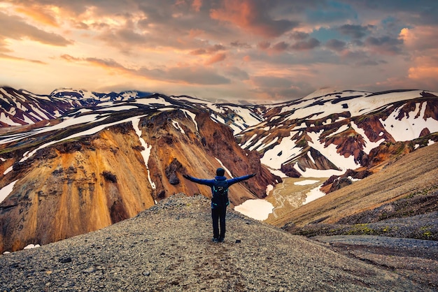 Hiker man standing with arms raised on the cliff with volcanic mountain and snow covered from Blanhjukur trail on summer in the evening