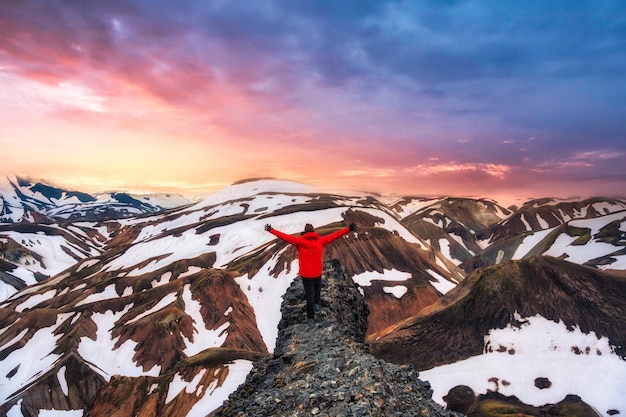 火山の山と雪に覆われた崖の上に腕を上げて立っているハイカーの男は、ランドマンナラウガルで夏に景色を覆う