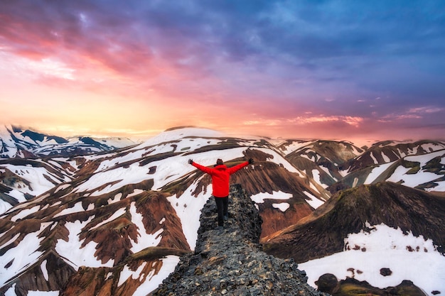 란트만날라우가르(Landmannalaugar)에서 여름에 화산 산과 눈 덮인 전망이 있는 절벽에 팔을 들고 서 있는 등산객