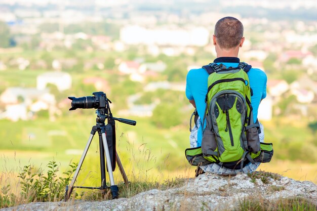 Hiker фотограф, сидя на скале, с нетерпением жду возле камеры на штативе