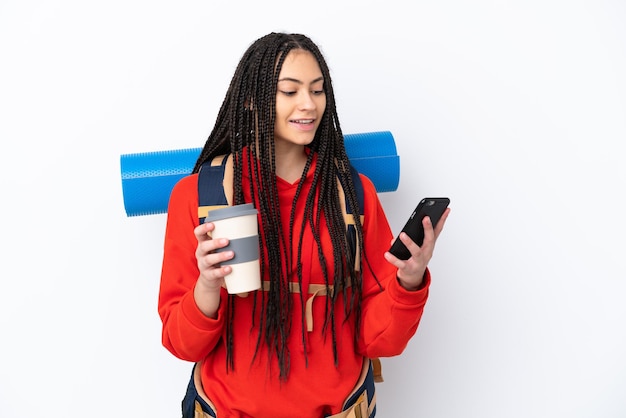 Hiker teenager girl with braids over isolated white background holding coffee to take away and a mobile