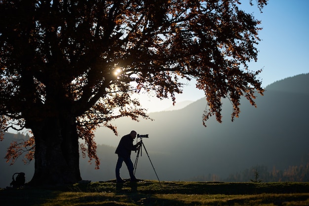 Hiker tourist man with camera on grassy valley on background of mountain landscape under big tree.