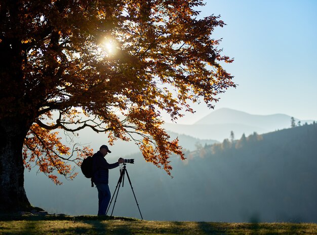 Hiker tourist man with camera on grassy valley on background of mountain landscape under big tree.