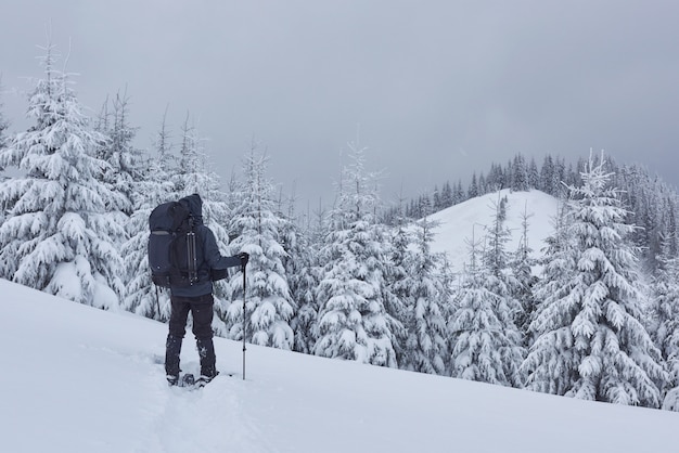 Hiker, with backpack, is climbing on the mountain range, and admires snow-capped peak. Epic adventure in the winter wilderness