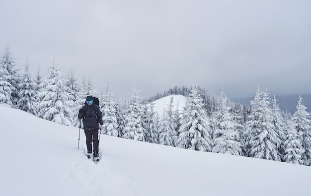 Hiker, with backpack, is climbing on the mountain range, and admires snow-capped peak. Epic adventure in the winter wilderness