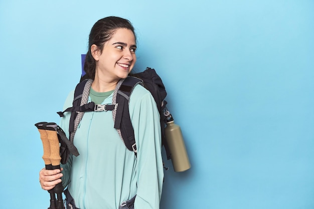 Photo hiker with backpack and poles on blue looks aside smiling cheerful and pleasant