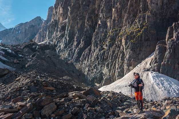 Hiker with trekking poles stands on moraine against rocks with glacier in bright sunlight Man in sunglasses in bright sun near sunlit rocky mountain wall Tourist in high mountains in sunny day