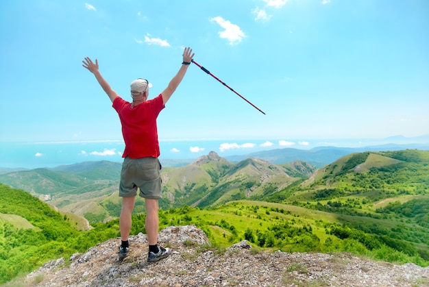 Hiker young man with trekking pole standing on cliff and looking at green mountains