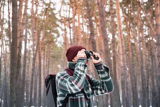 写真を撮る冬の森で男性の人をハイキングします。美しい雪に覆われた森の中で市松模様の冬シャツの男は、古いフィルムカメラを使用します