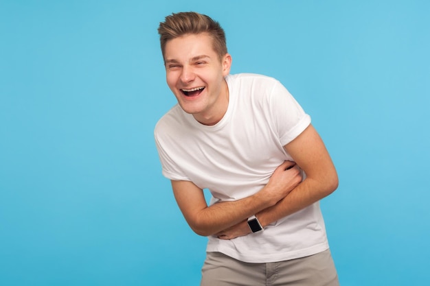 Hilarious joke Portrait of laughing man in casual white tshirt holding his stomach and hunched in crazy hysterical laughter sincere joyful emotions indoor studio shot isolated on blue background