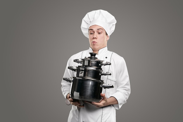 Hilarious male chef in white uniform and with stack of saucepans standing on gray background in studio