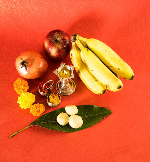 Photo hindu puja elements, diya, haldi kumkum,apple, pomegranate and banana as offering to god and indian sweet pedha in silver bowl top view, isolated on red surface