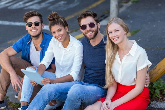 Hip friends holding tablet and sitting on sidewalk in the city