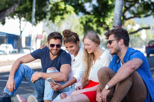 Hip friends looking at smartphone and sitting on sidewalk in the city