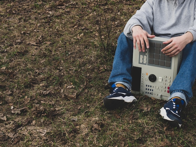 A hiphop beatmaker sits on the grass in a park under a tree holding an oldfashioned ' 90s drum machine Retro Digital Musical Instrument for Hip Hop Producers and Beatmakers