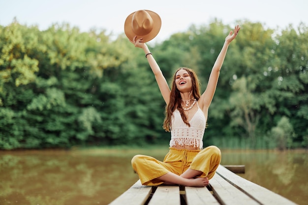 Foto hippie ecoattivista donna viaggiatrice si siede su un ponte vicino a un lago con le braccia stese con un cappello e sorridendo sinceramente foto di alta qualità