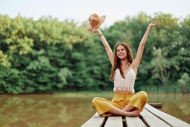 Photo hippie ecoactivist woman traveler sits on a bridge by a lake with her arms outstretched with a hat and smiling sincerely high quality photo