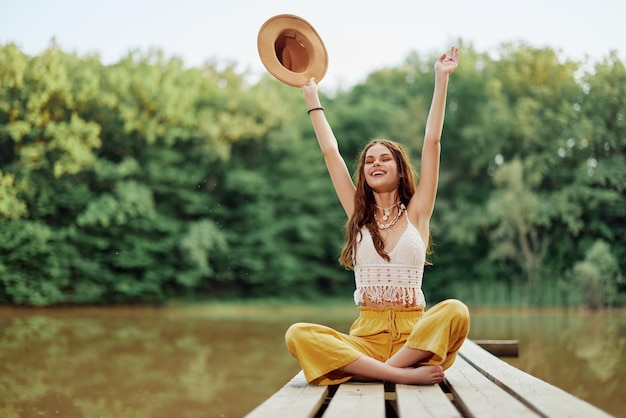 Hippie ecoactivistische vrouwelijke reiziger zit op een brug bij een meer met haar armen uitgestrekt met een hoed en oprecht glimlachend