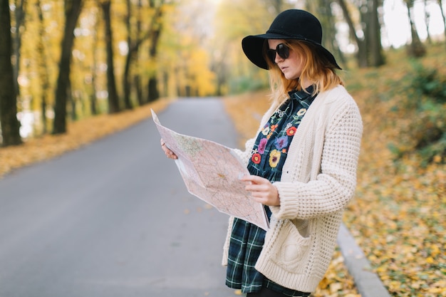 Ragazza del hippie con una mappa, vecchia macchina fotografica in un maglione lavorato a maglia, occhiali da sole e cappello cammina parco autunnale.