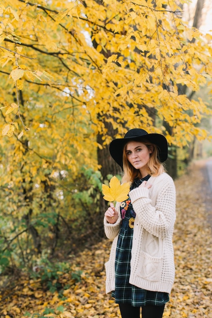 Hippie meisje met een gele maple leaf in een gebreide trui en hoed wandelingen herfst park.