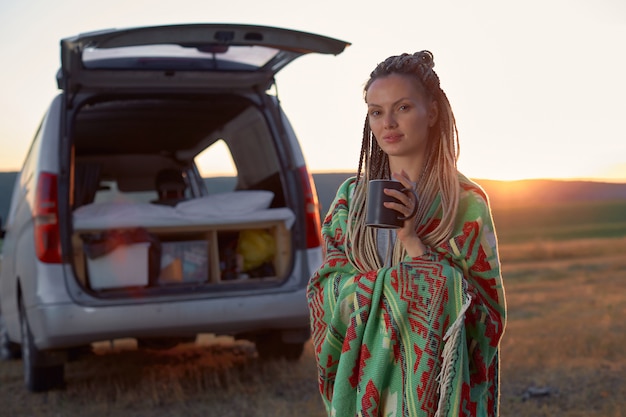 A hippie woman in a bright ethnic blanket holds a mug standing in a field at sunset near her car hig...