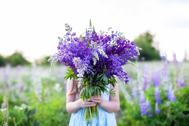 野生の花の花束を手に持ったヒッピーの女の子。女の子はルピナスの花束の後ろに顔を隠しました。小さな女の子は、開花フィールドに紫のルピナスの大きな花束を保持しています。自然のコンセプト。プレゼント
