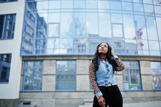 Hipster african american girl wearing jeans shirt with leopard sleeves posing at street against modern office building with blue windows