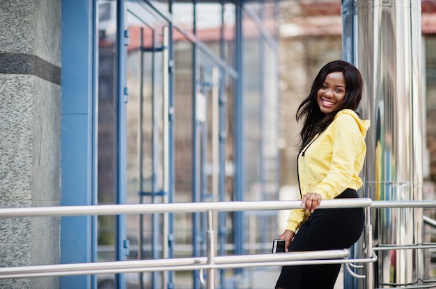 Hipster african american girl wearing yellow hoodie posing at street against office building with blue windows.