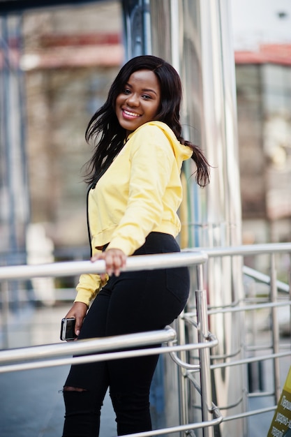 Hipster african american girl wearing yellow hoodie posing at street against office building with blue windows.