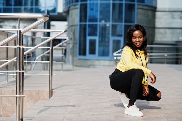 Hipster african american girl wearing yellow hoodie posing at street against office building with blue windows.